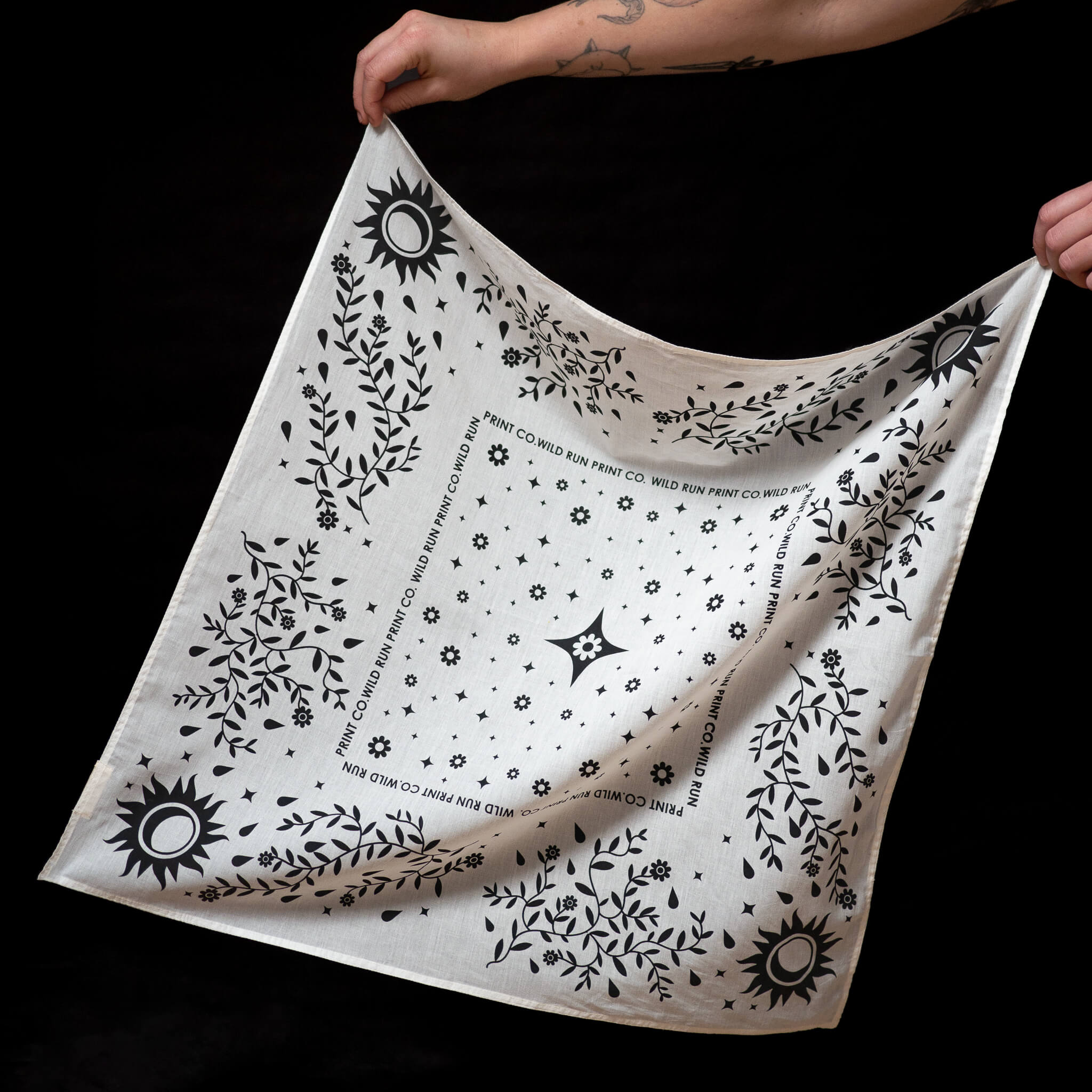 A person holds up a Wild Run Print Co. bandana with black and white botanical sun and flower designs against a black background