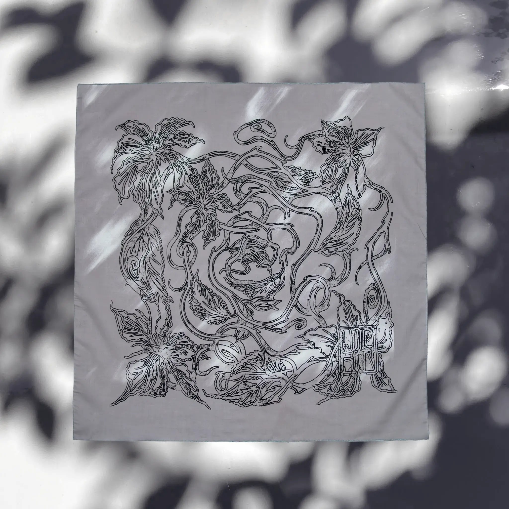 A square gray scarf featuring an intricate black line-art illustration of flowers and leaves, displayed flat against a background with soft leaf shadows.