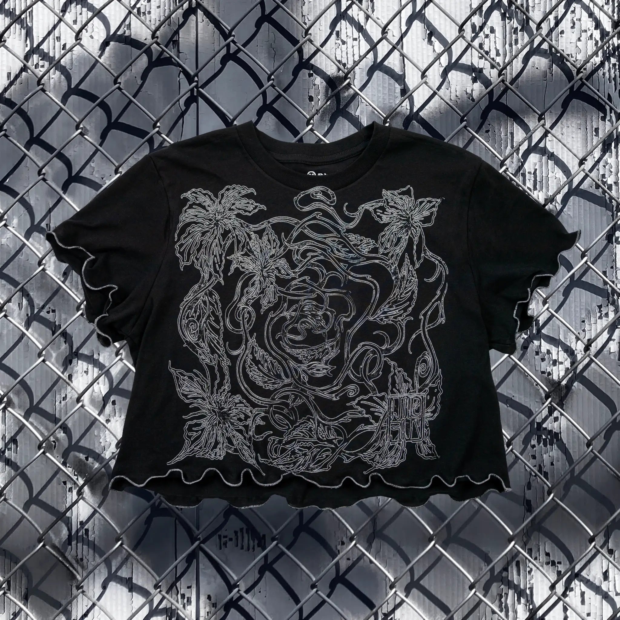 A black cropped t-shirt with ruffled sleeves and hem, featuring a large white line-art graphic of vines and leaves, laid flat against a chain-link fence background.